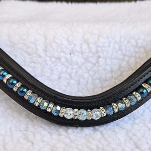 May include: A black leather horse bridle browband with a curved design. The browband is decorated with alternating rows of clear and blue crystal beads and silver accents. The browband is designed for a horse.