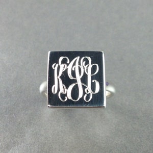 May include: A silver ring with a square top featuring a monogram engraved in a cursive font.