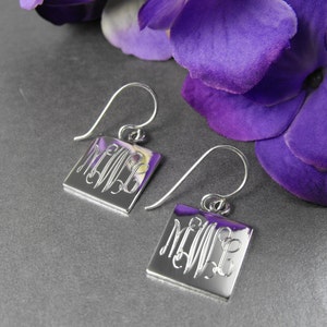 May include: A pair of silver square earrings with a monogram design. The earrings are hanging from a silver hook.