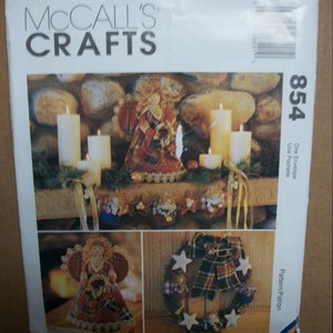 May include: McCall's Crafts pattern envelope featuring a Christmas theme. The image showcases a decorated mantel with candles, an angel figurine, and a wreath with stars and plaid ribbon. The number 854 is visible on the envelope.
