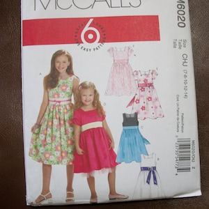 May include: A McCall's sewing pattern for girls' dresses, sizes 7-8, 10-12, and 14. The pattern includes four different dress styles: a floral print dress, a pink dress with a white sash, a blue dress with a black sash, and a white dress with a purple sash. The pattern is labeled "M6020/CHJ 2".