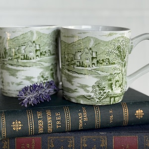 May include: Two white ceramic mugs featuring a green transferware-style landscape print of houses, trees, and hills. The mugs have a cylindrical shape with a curved handle on the right side. A small sprig of purple lavender rests in front of the mugs.