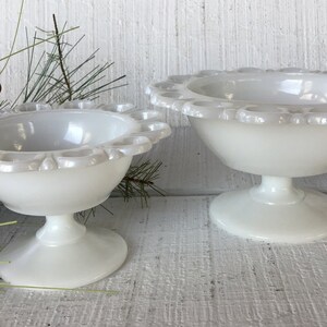 May include: Two white milk glass pedestal bowls with scalloped edges. The larger bowl is in the background and the smaller bowl is in the foreground.
