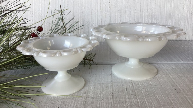 May include: Two white milk glass pedestal bowls with scalloped edges. The larger bowl is in the background and the smaller bowl is in the foreground.