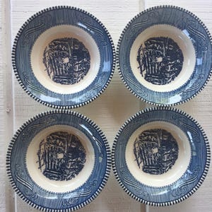 May include: A set of four round ceramic bowls featuring a cream-colored center with a dark blue transferware scene of a rural cottage. The wide rims are decorated with a dark blue hatched pattern, scrollwork, and a beaded edge detail.