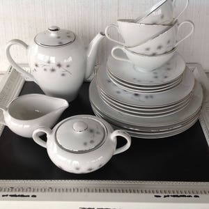 May include: A white tea set with a floral design. The set includes a teapot, creamer, sugar bowl, and four cups and saucers. The set is displayed on a white tray with a black surface.