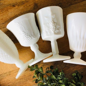 May include: Four white milk glass goblets with different embossed patterns. The goblets are on a wooden surface.