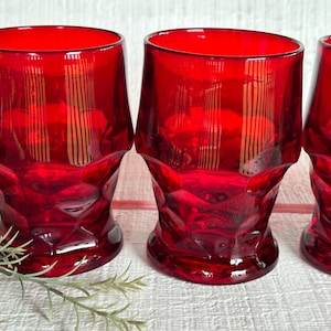 May include: Three red glass tumblers with a faceted design. The glasses have a gold-colored stripe pattern.