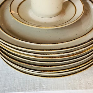 Może przedstawiać: Stos beżowych ceramicznych talerzy obiadowych z brązową obw&oacute;dką. Talerze są ułożone w okrągły wz&oacute;r, przy czym g&oacute;rny talerz jest największy, a dolny najmniejszy.