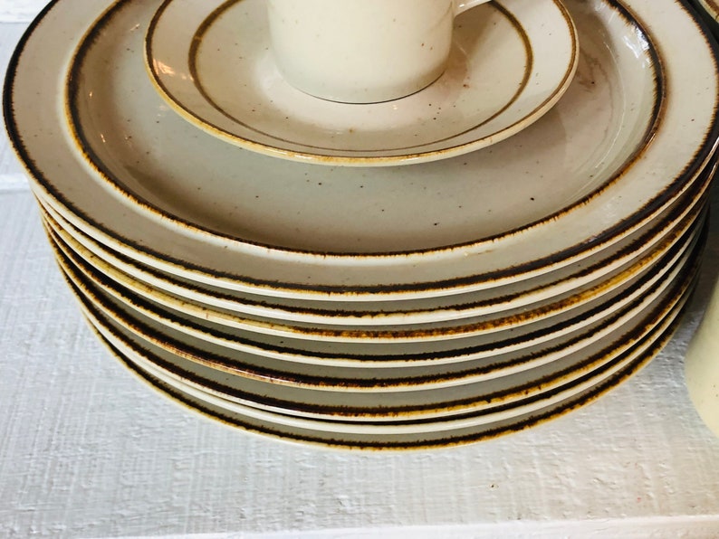 Może przedstawiać: Stos beżowych ceramicznych talerzy obiadowych z brązową obw&oacute;dką. Talerze są ułożone w okrągły wz&oacute;r, przy czym g&oacute;rny talerz jest największy, a dolny najmniejszy.
