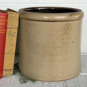 May include: A cylindrical stoneware crock with a speckled tan finish and a dark brown rim. Beside the crock are three vintage hardcover books with red and tan spines, featuring titles including Don Lorenzo's Bride and The Pillar of Fire.