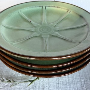 May include: A stack of four round ceramic plates featuring a light sage green glaze on the interior surface with a raised, radiating sunburst pattern. The edges and undersides of the plates are finished in a contrasting dark brown glaze.
