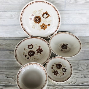 May include: Set of five white ceramic plates with brown floral designs. The plates include a large dinner plate, two medium-sized bowls, and two smaller bowls.