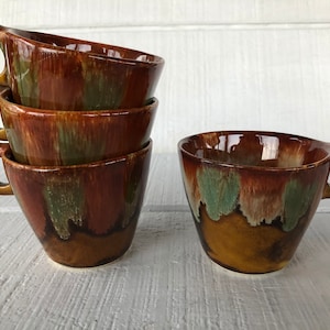 May include: A set of five ceramic coffee cups. The cups have a unique design with a brown, green, and tan glaze. Each cup has a handle with a circular opening. The cups are stacked and arranged on a white surface.