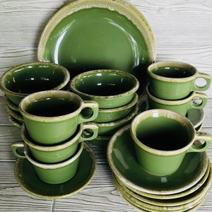 May include: A green ceramic dinnerware set with a cream-colored rim. The set includes plates, bowls, saucers, and teacups. The teacups are stacked, and the plates and bowls are arranged in a neat display. The set is ideal for a kitchen or dining room.