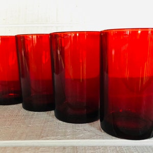 May include: Four red-tinted drinking glasses. The glasses are cylindrical with a slightly tapered shape. The color is a deep, rich red, and the glasses are arranged in a row on a white surface.