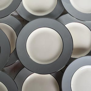 May include: A collection of round dinner plates arranged in an overlapping pattern. Each plate features a smooth, cream-colored center surrounded by a wide, solid matte gray rim. The design is consistent across all plates, showing a circular shape.