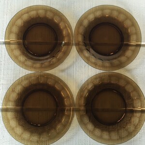 以下が含まれることがあります： テクスチャ加工されたスカラップリムデザインの、4つの茶色のガラスプレート。白い木製の表面に配置され、円形と色を際立たせています。これらのヴィンテージプレートは、食品の提供に適しています。