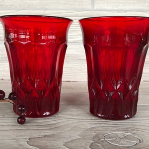 May include: Two red glass tumblers with a diamond pattern. The glasses are on a white wooden surface.