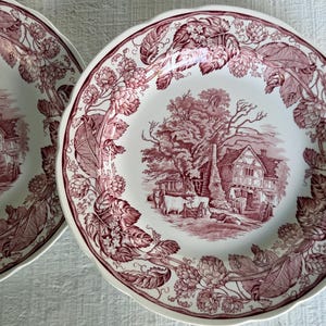 May include: Two white ceramic plates featuring a maroon transferware pattern. The center depicts a rural scene with a timber-framed house and cattle, while the scalloped rim is decorated with a detailed border of hop vines and leaves.