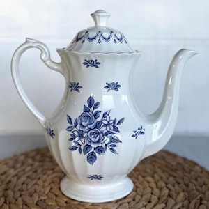 May include: A white porcelain teapot featuring a blue floral rose pattern and a fluted body design. The teapot includes a matching lid with a blue decorative border, a curved handle, and a long spout, all decorated with small blue floral accents.
