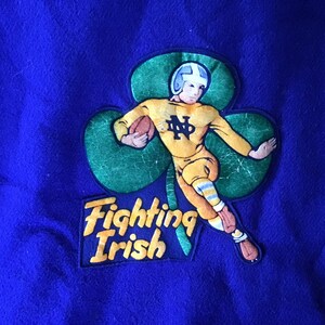 K&ouml;nnte beinhalten: Ein blauer Filz-Patch mit einem gr&uuml;nen Kleeblatt und einem Cartoon-Footballspieler in einem gelben Trikot mit den Buchstaben "ND" auf der Vorderseite. Der Spieler h&auml;lt einen Football und rennt. Der Text "Fighting Irish" befindet sich unter dem Kleeblatt.