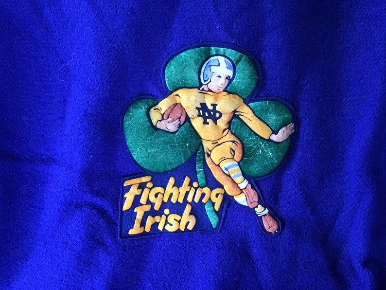 K&ouml;nnte beinhalten: Ein blauer Filz-Patch mit einem gr&uuml;nen Kleeblatt und einem Cartoon-Footballspieler in einem gelben Trikot mit den Buchstaben "ND" auf der Vorderseite. Der Spieler h&auml;lt einen Football und rennt. Der Text "Fighting Irish" befindet sich unter dem Kleeblatt.