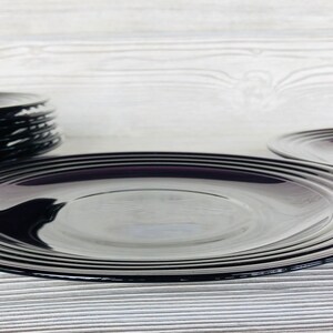 May include: A set of black glass plates with a subtle purple hue. The plates are stacked on top of each other, with the top plate being the largest. The plates are on a white wooden surface.