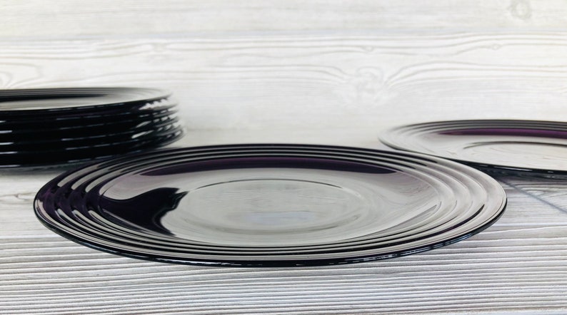 May include: A set of black glass plates with a subtle purple hue. The plates are stacked on top of each other, with the top plate being the largest. The plates are on a white wooden surface.