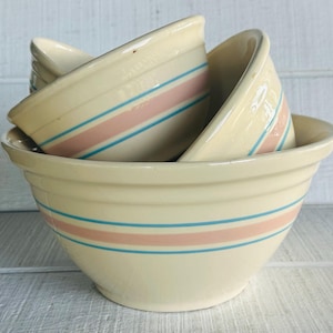 May include: A set of cream-colored ceramic mixing bowls stacked together. Each bowl features a wide horizontal band of soft pink flanked by two thinner horizontal bands of light blue. The bowls have a ribbed exterior texture and a smooth finish.