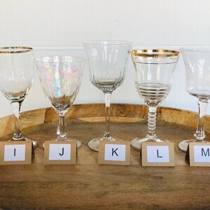 May include: Five clear glass wine glasses with gold rims on a wooden tray. The glasses are labeled with letters I, J, K, L, and M.