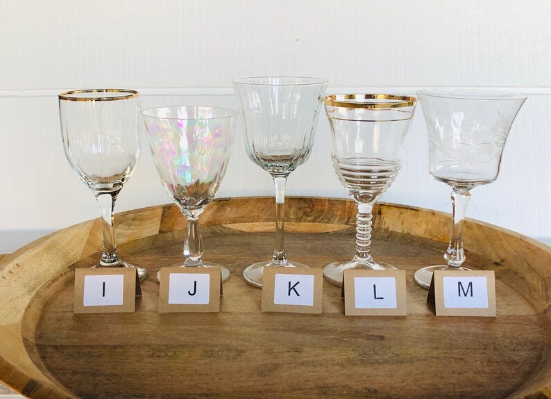 May include: Five clear glass wine glasses with gold rims on a wooden tray. The glasses are labeled with letters I, J, K, L, and M.