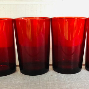 May include: Four red drinking glasses. The glasses are made of a deep red glass that fades to a darker shade at the base. The glasses are cylindrical with a slight taper and are evenly spaced.