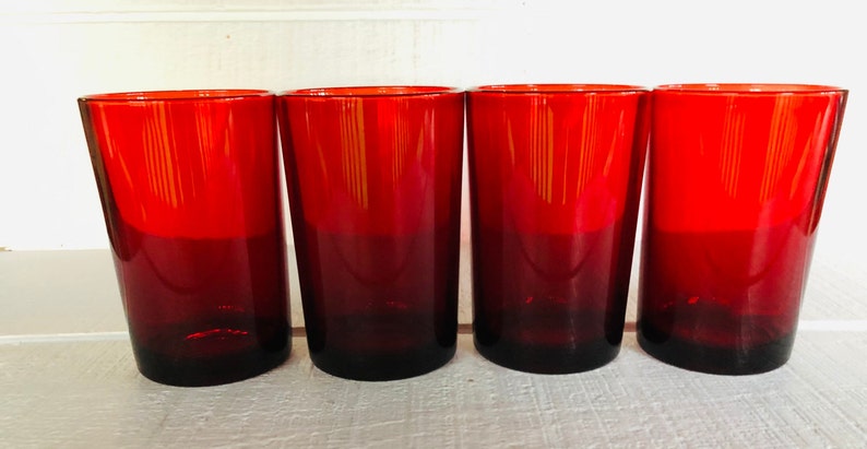 May include: Four red drinking glasses. The glasses are made of a deep red glass that fades to a darker shade at the base. The glasses are cylindrical with a slight taper and are evenly spaced.