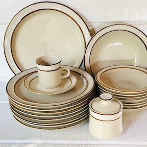 May include: A set of brown and white ceramic dinnerware with a brown rim. The set includes a large dinner plate, a smaller plate, a bowl, a cup, and a sugar bowl.