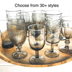 May include: A collection of various grey-tinted glass goblets and tumblers with different textures, including ribbed and embossed patterns, arranged together. The text "Choose from 30+ styles" is displayed above the glassware.