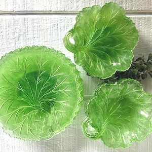 May include: Three green glass serving dishes in the shape of leaves. The dishes have a textured surface with detailed vein patterns. One is round, and two are leaf-shaped with handles. The dishes are arranged on a white surface.