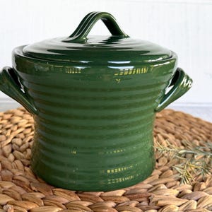 May include: A dark green ceramic lidded pot with a glossy finish and horizontal ribbed texture. The vessel features two side handles and a matching lid with a central arched handle, showcasing a uniform deep forest green color throughout.