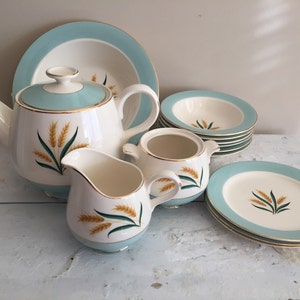 May include: A vintage tea set featuring a white teapot, creamer, sugar bowl, plates, and bowls. Each piece has a light blue border and gold trim, with a wheat stalk design. The set is arranged on a white surface.