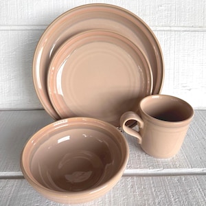 May include: A set of beige ceramic dinnerware, including a large plate, a smaller plate, a bowl, and a mug. The plates and bowl have a subtle, darker rim. The mug has a curved handle.