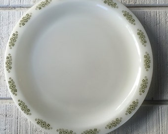 May include: White plate with a floral design around the rim. The plate has a simple, elegant design with small, dark green flowers. The plate is round and appears to be made of ceramic or glass.