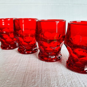 May include: Four red glass tumblers with a faceted design. The glasses are arranged in a row on a white surface.
