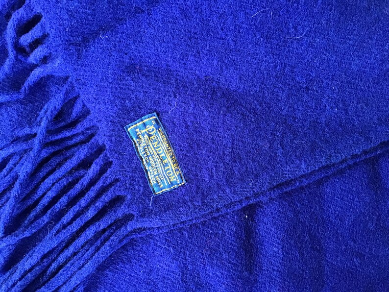 K&ouml;nnte beinhalten: Ein k&ouml;nigsblauer Pendleton Wollschal mit Fransen an den Enden. Der Schal hat ein blaues Etikett mit dem Pendleton-Logo und dem Aufdruck "WARRANTED TO BE A PENDLETON WOOL PRODUCT".