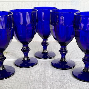 May include: Six cobalt blue wine glasses with faceted bowls and ornate stems. The glasses are arranged on a white surface, showcasing their deep color and elegant design. The stemware is ideal for serving wine.