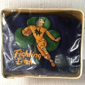 K&ouml;nnte beinhalten: Ein Vintage-Kissen der Notre Dame Fighting Irish in einem durchsichtigen Plastik-Etui. Das Kissen ist blau mit einem gr&uuml;nen Kleeblatt und einem Cartoon-Footballspieler in gelber Uniform. Der Text "Fighting Irish" ist auf das Kissen gedruckt.