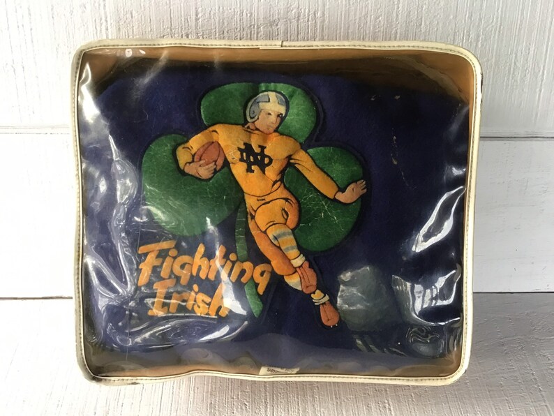 K&ouml;nnte beinhalten: Ein Vintage-Kissen der Notre Dame Fighting Irish in einem durchsichtigen Plastik-Etui. Das Kissen ist blau mit einem gr&uuml;nen Kleeblatt und einem Cartoon-Footballspieler in gelber Uniform. Der Text "Fighting Irish" ist auf das Kissen gedruckt.