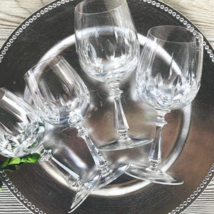May include: Five clear crystal wine glasses with intricate etched designs are arranged on a silver platter with a beaded edge. The glasses are positioned at various angles, showcasing their elegant stems and bowls. A sprig of green foliage adds a touch of color.