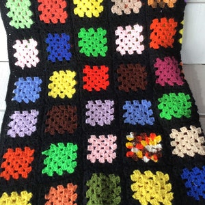 May include: A handmade granny square blanket with a black background. The squares are in various colors, including red, yellow, green, blue, and purple. The blanket is made of yarn and has a textured appearance. The blanket is a cozy home decor item.
