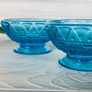 May include: Two blue glass bowls with a textured design and a pedestal base. The bowls are on a white wooden surface.