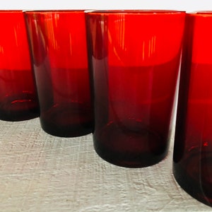 May include: Four tall, red-tinted drinking glasses. The glasses have a simple, cylindrical shape with a slightly tapered design. The color is a deep, rich red, and the glasses appear to be made of glass. The glasses are arranged in a row.